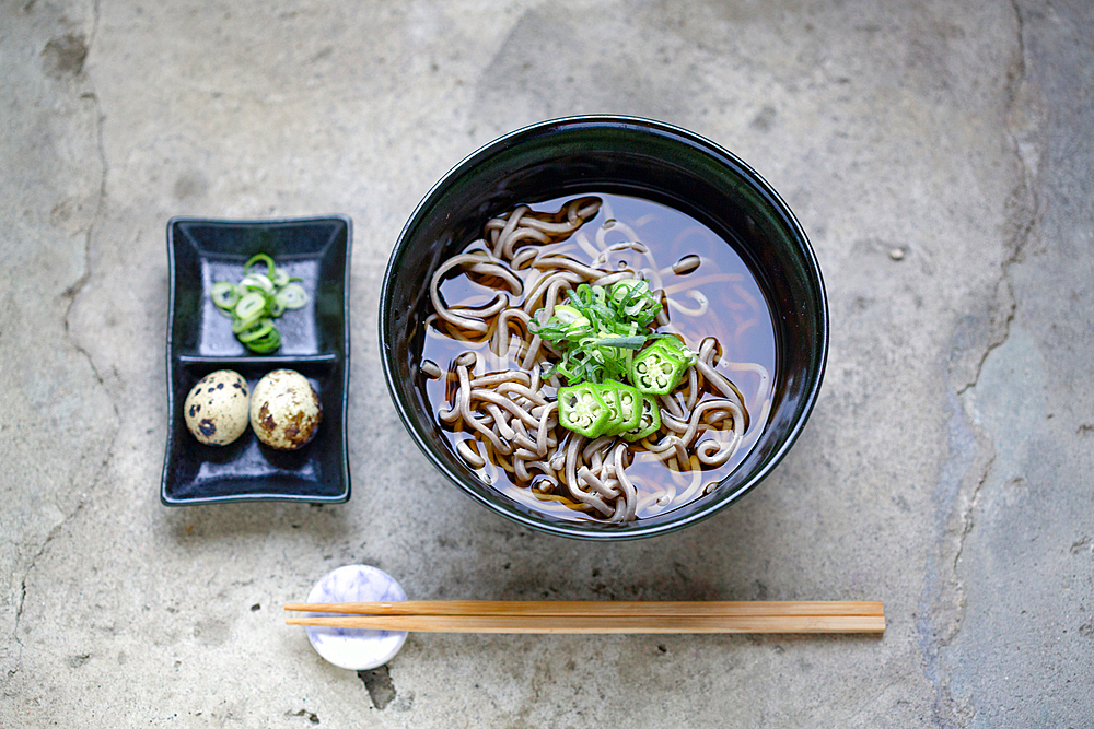 A black china bowl of noodles and broth, sliced green chilli and a dish with quail's eggs and sliced vegetables.