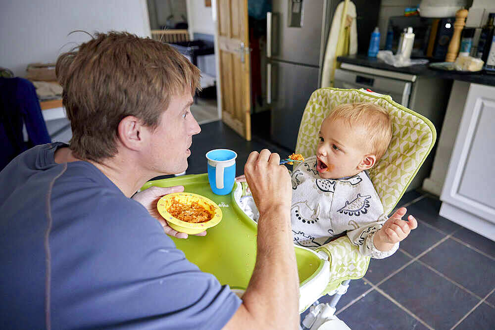 Father feeding toddler in high chair in kitchen