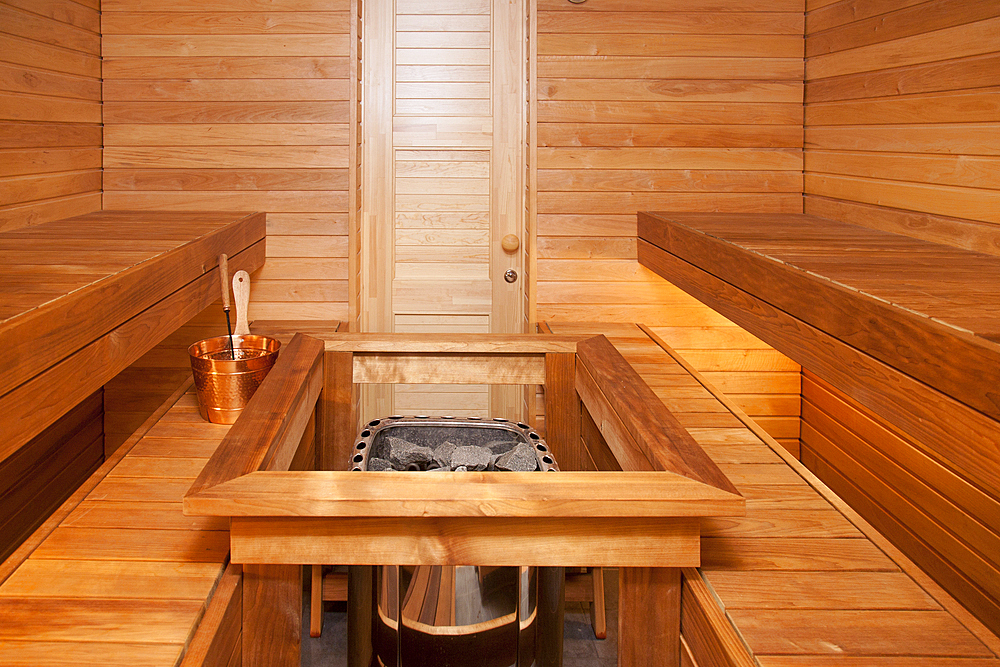 Interior of a wooden panelled steam room, sauna, with central stove or hot stone, a basket and ladle for water.