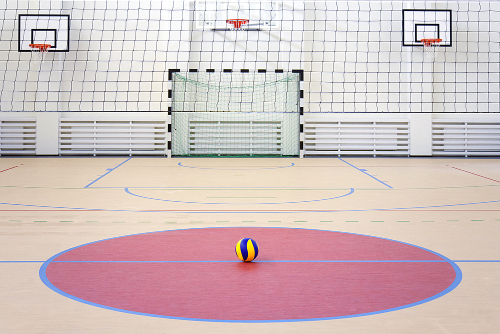 A school sports hall with a marked indoor football pitch, goal and basketball hoops, a striped blue and yellow ball in a pink circle.