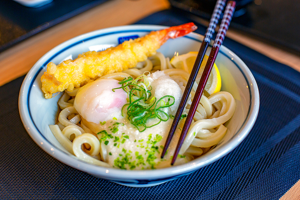 A dish of noodles, vegetables and fish and a tempura prawn, chopsticks.