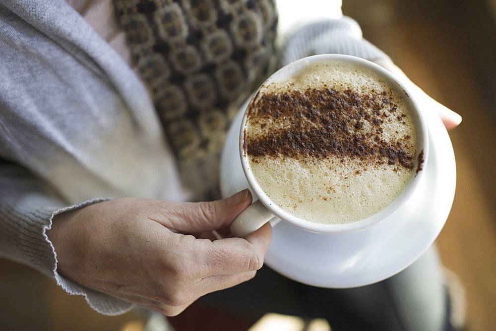 A person holding a full cup of frothy cappuccino coffee in a white china cup. Chocolate powder sprinkled in a pattern on the top. Coffee shop, Kingston, New York, USA