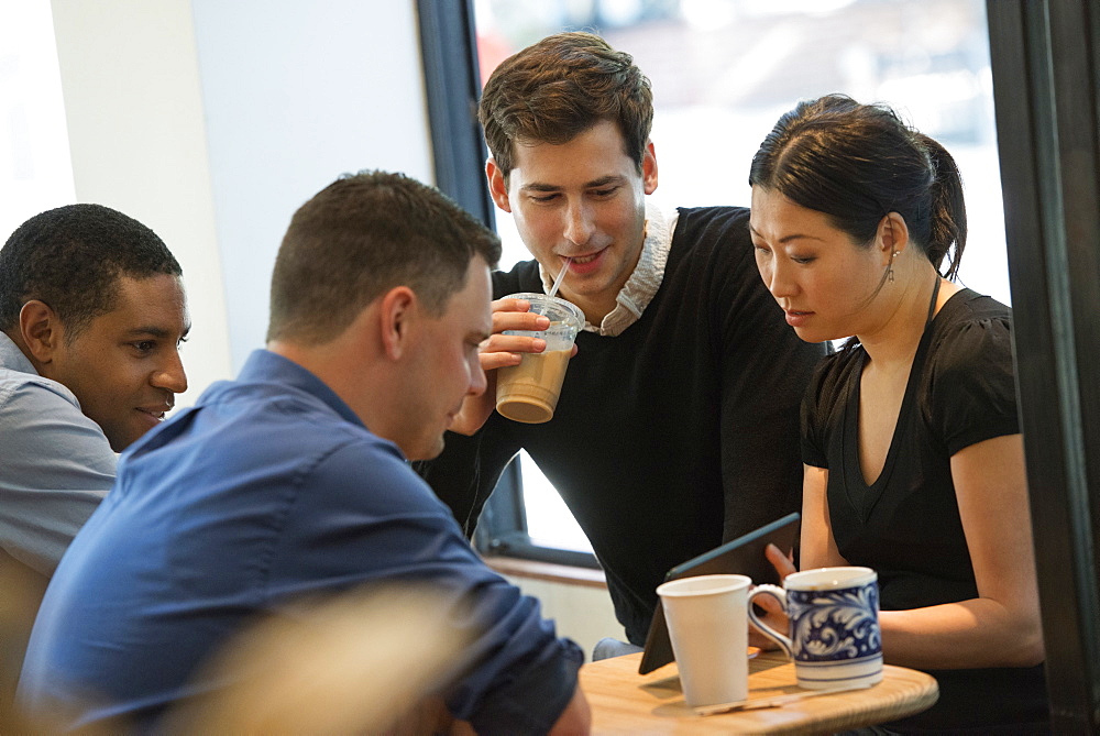 A group of people sitting around a table in a coffee shop. Looking at the screen of a digital tablet. Three men and a woman.