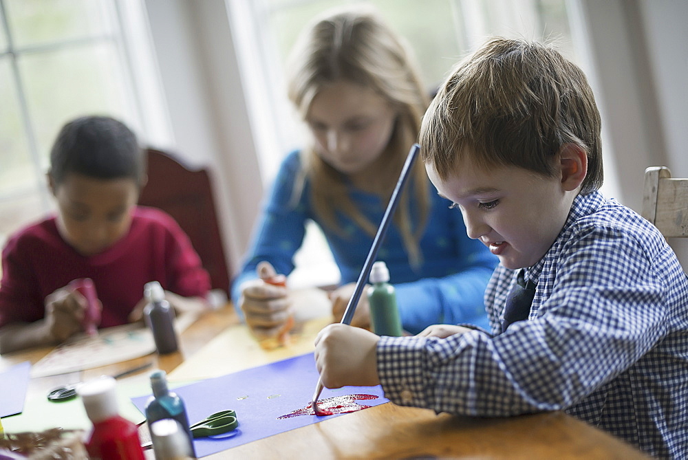 Children in a family home. Three children sitting at a table using glue and paint to create decorations, Woodland Valley, New York, USA