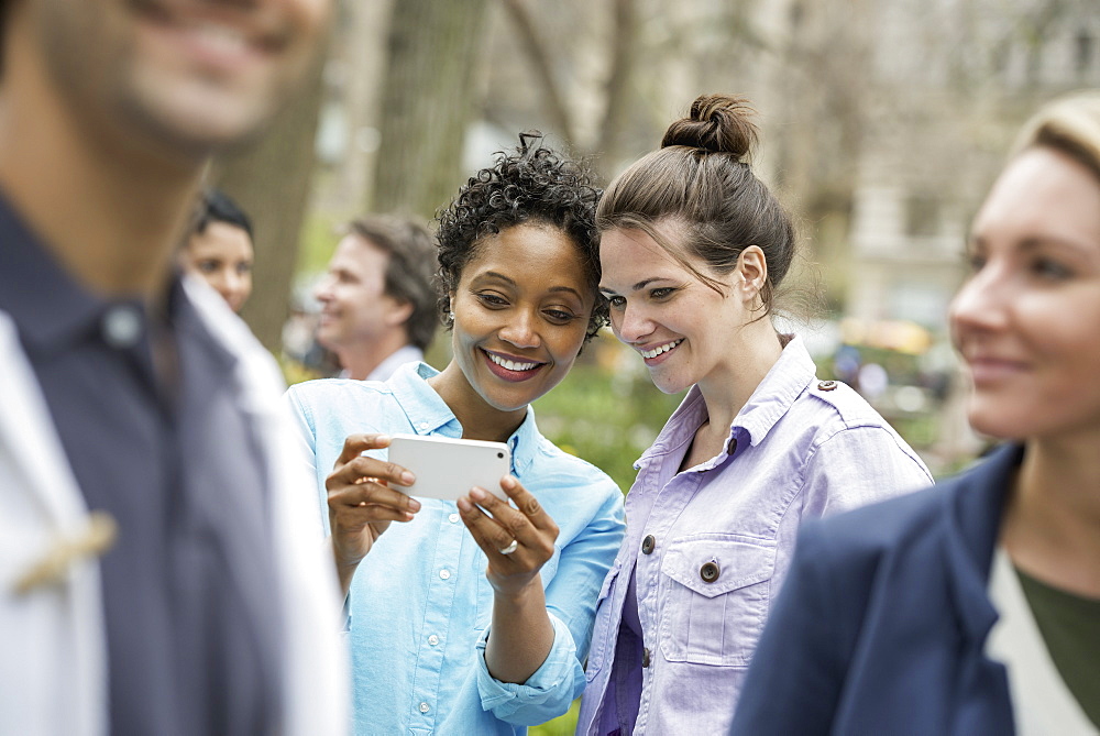 People outdoors in the city in spring time. New York City park. Two women in a group of friends, looking at a cell phone and smiling, New York city, USA