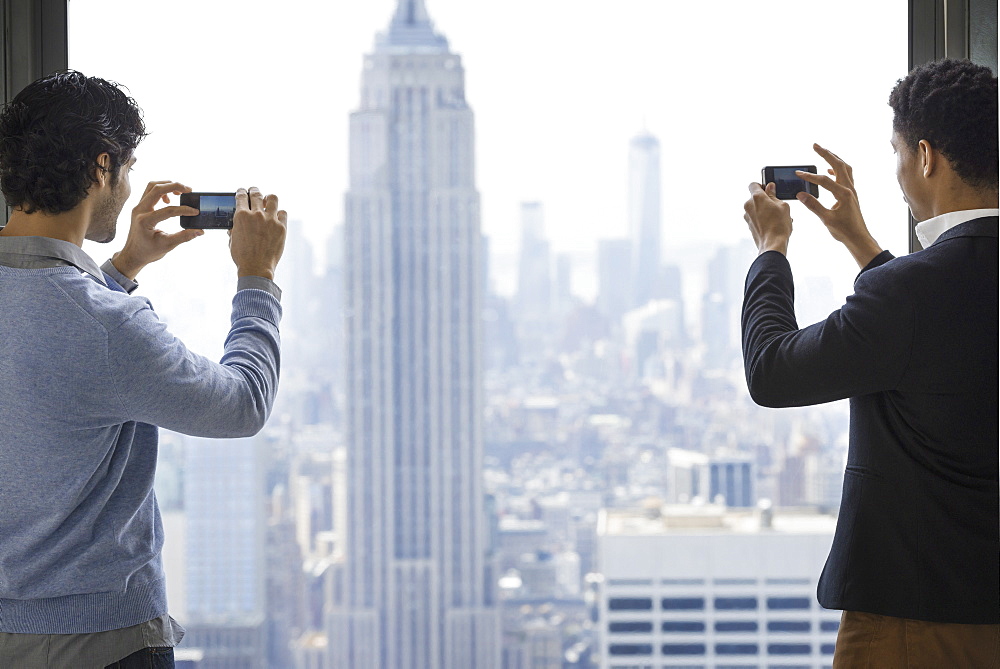 Urban lifestyle. Two young men using their phones to take images of the city from an observation platform overlooking the Empire State Building, New York city, USA