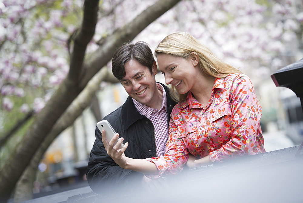 City life in spring. Young people outdoors in a city park. A young woman and man sitting side by side looking down at a smart phone, New York city, USA