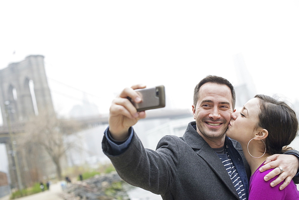 New York city. The Brooklyn Bridge crossing over the East River. A couple taking a picture with a phone, a selfy of themselves, New York city, USA