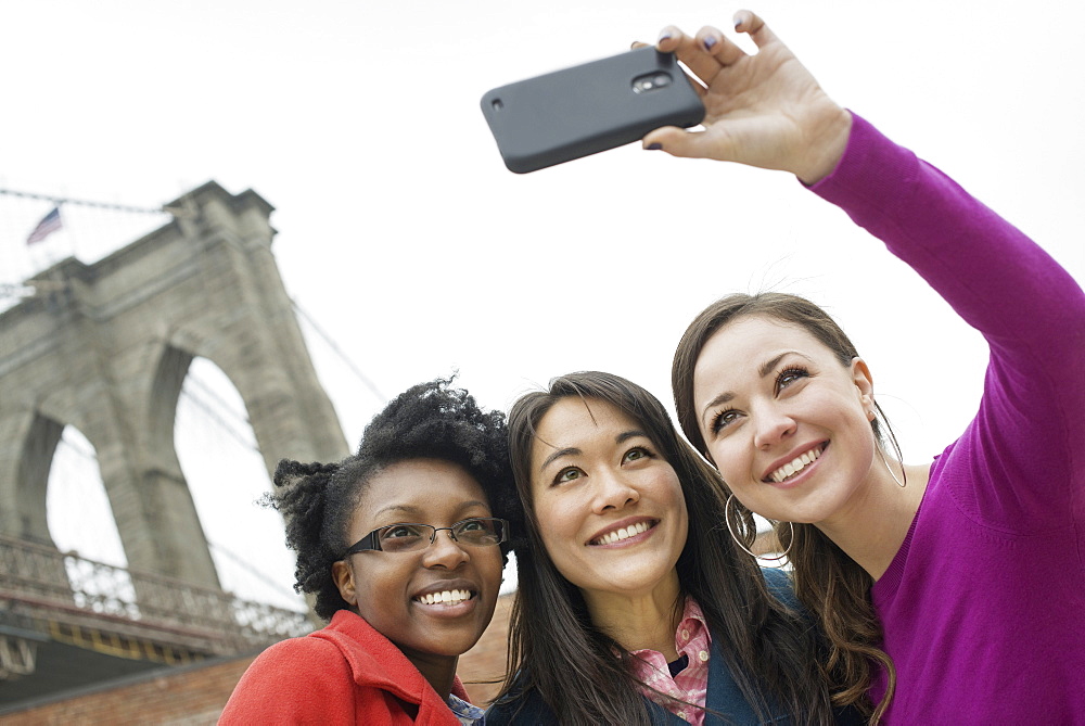 New York city, the Brooklyn Bridge crossing over the East River. Three women in a row, smiling, as one takes a picture with a smart phone, New York city, USA