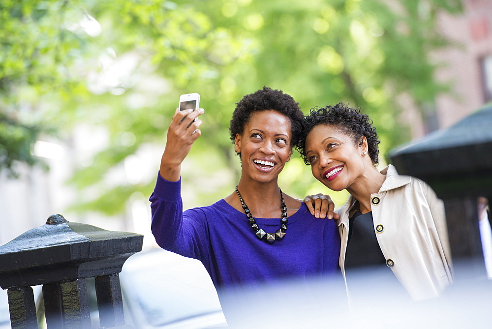 City life. Two women sitting on a park bench, side by side. Posing for a photograph with a smart phone. 