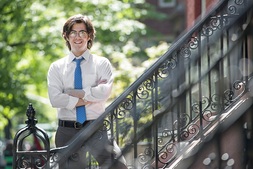 City life. People on the move. A young man in w white shirt and blue tie, standing with arms folded outside a townhouse, on the steps. 