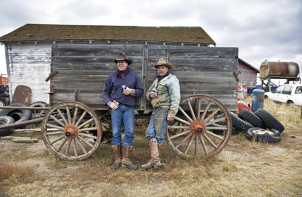 Two men in cowboy hats and cowboy boots leaning against a wooden wagonCowboys, Saskatchewan, Canada