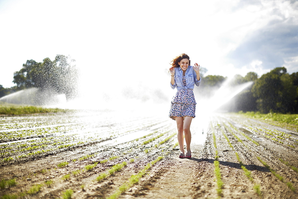 A young woman in denim jacket standing in a field, irrigation sprinklers working in the background, New York State, USA