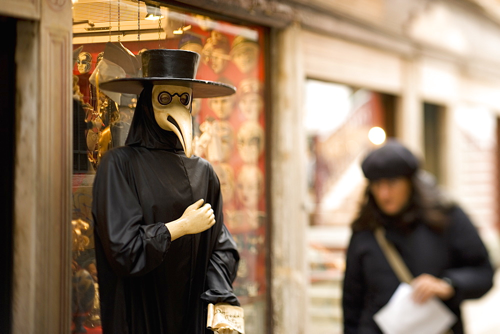 A person in a bird mask, a traditional carnival costume, with a long bird beak, A woman walking by, Venice, Italy