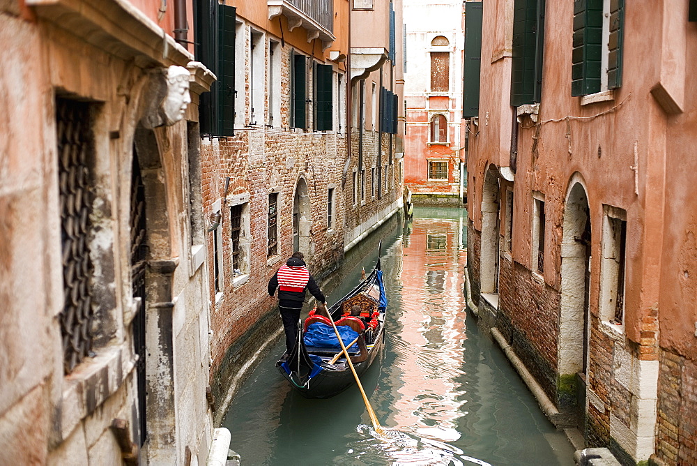 A gondola boat gliding down a small narrow waterway, between historic houses in the city of Venice, Venice, Italy