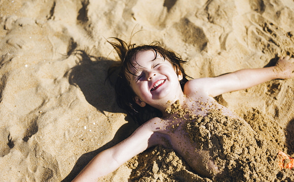 A boy lying on his back on the beach, his torso covered in sand, laughing, England