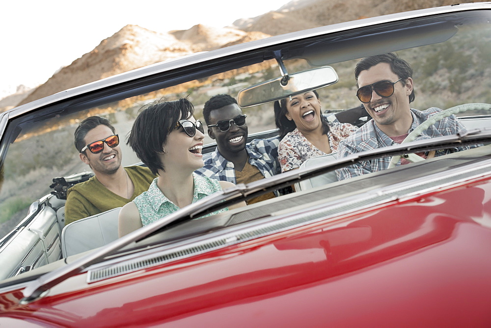 A group of friends in a red open top convertible classic car on a road trip, United States of America