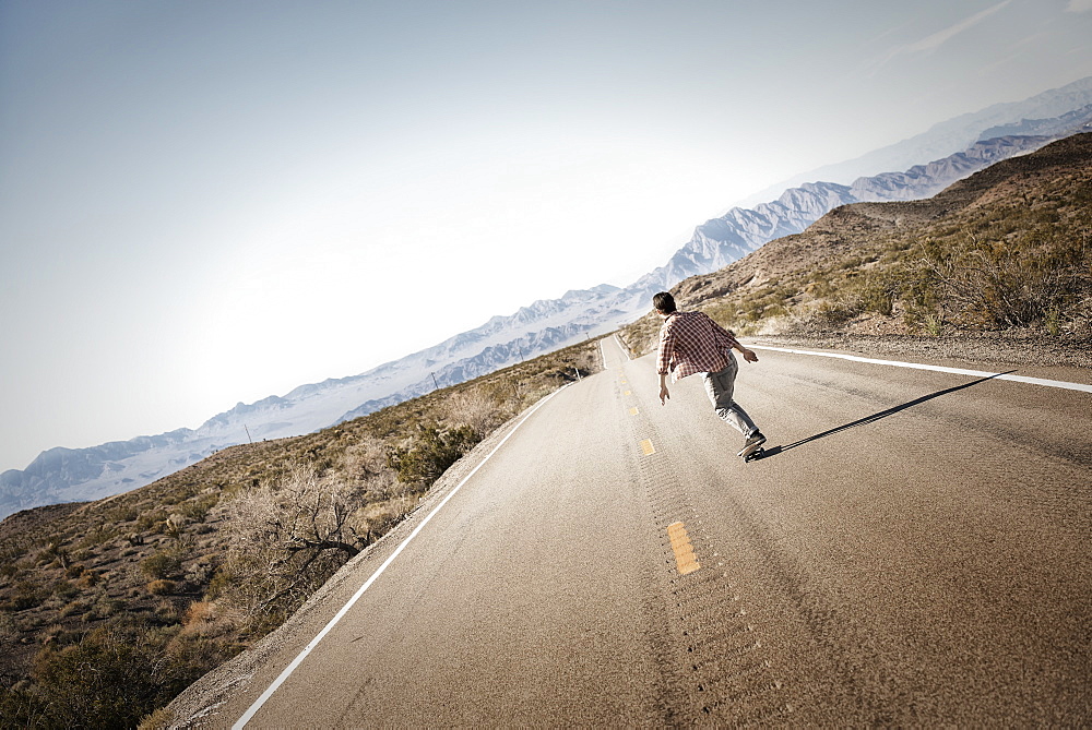 A young man riding down a tarmac road in the desert on a skateboard, United States of America