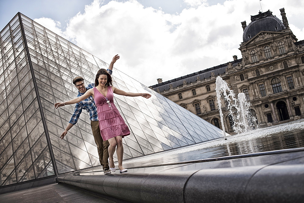 A couple in the courtyard of the Louvre museum, by the large glass pyramid. Water jets and shallow pool, France