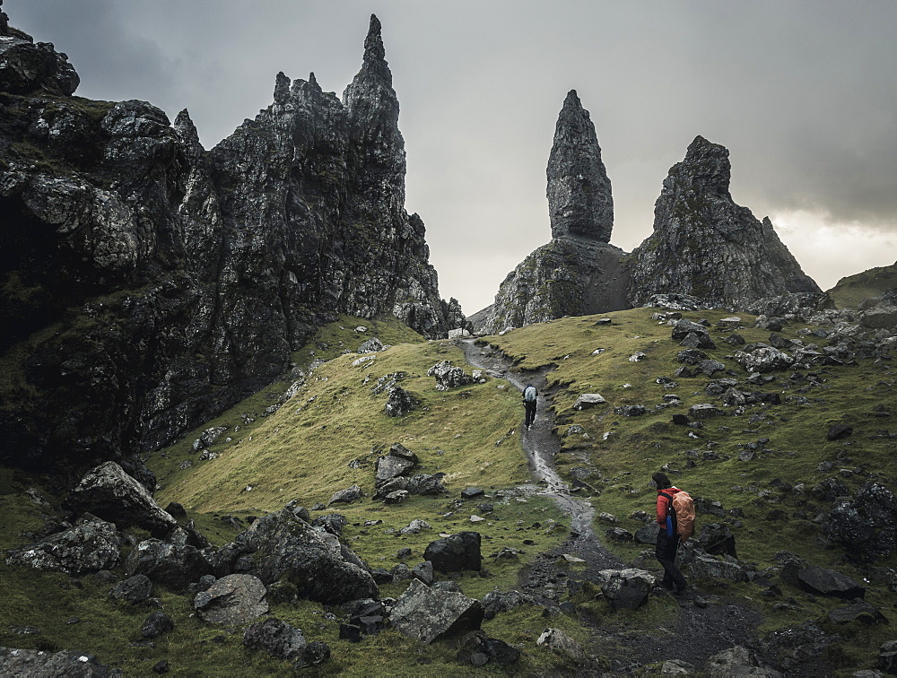 Two people with rucksacks on a narrow path rising to a dramatic landscape of rock pinnacles on the skyline towering above them, under an overcast sky with low cloud, Scotland, United Kingdom