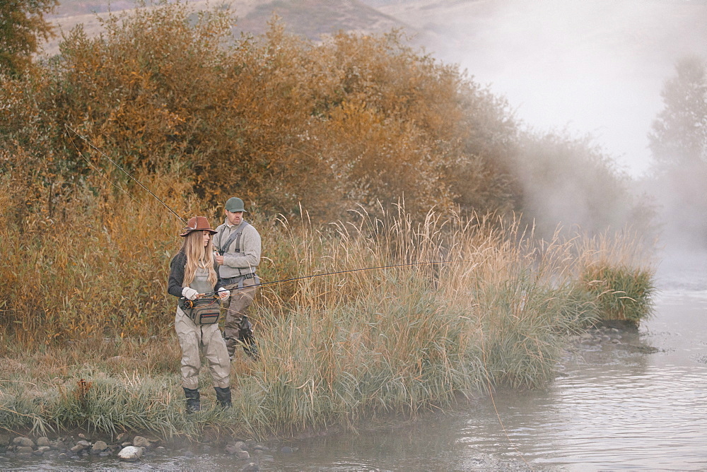 A man and woman standing on a riverbank, fishing.