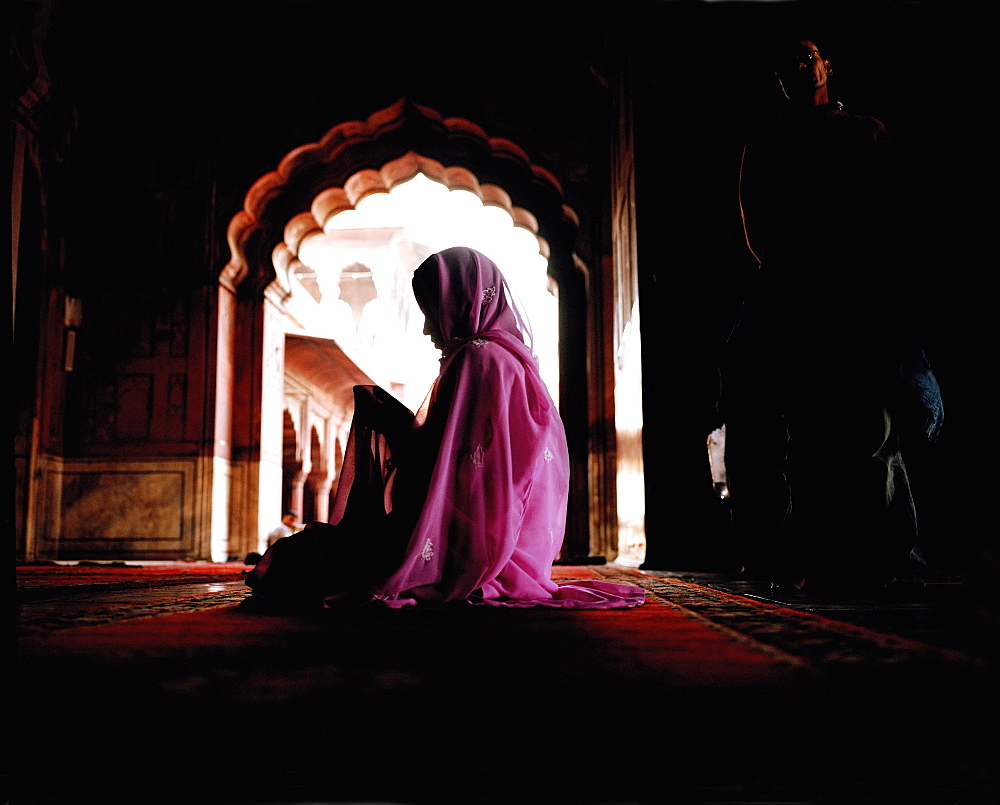 Woman wearing a pink veil and sari sitting on the floor covered with carpet. Scalloped archway. Deep shadow, India