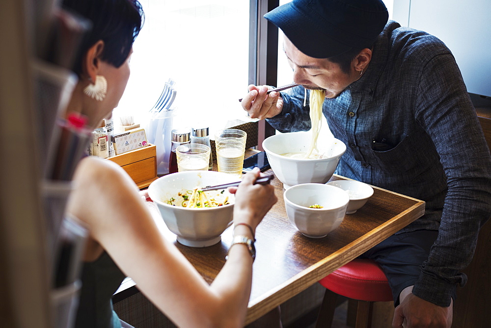 A ramen noodle cafe in a city. A man and woman seated eating noodles from large white bowls, Japan