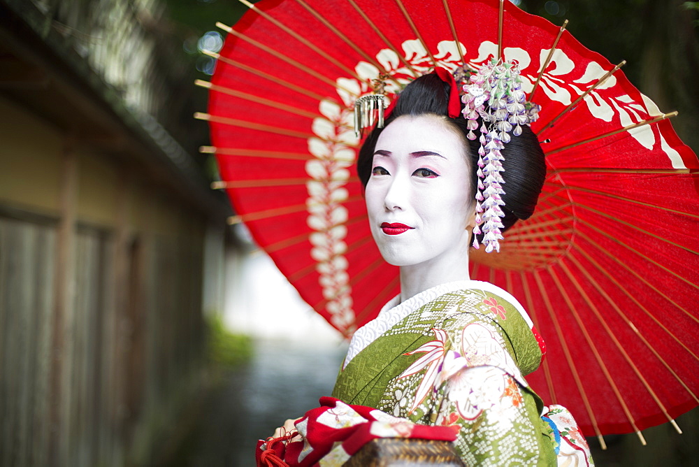 A woman dressed in the traditional geisha style, wearing a kimono and obi, with an elaborate hairstyle and floral hair clips, with white face makeup with bright red lips and dark eyes holding a red paper parasol, Japan