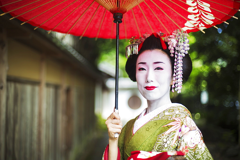 A woman dressed in the traditional geisha style, wearing a kimono and obi, with an elaborate hairstyle and floral hair clips, with white face makeup with bright red lips and dark eyes holding a red paper parasol, Japan