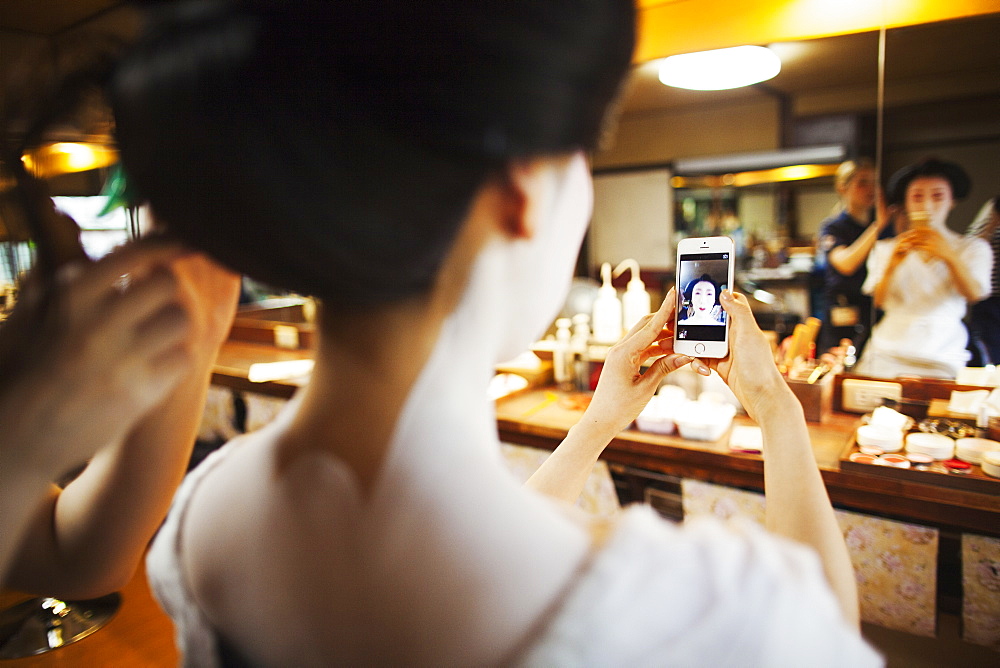 A geisha or maiko with a hair and make up artist creating the traditional hair style and make up. Woman making a selfie in the mirror with a smart phone, Japan