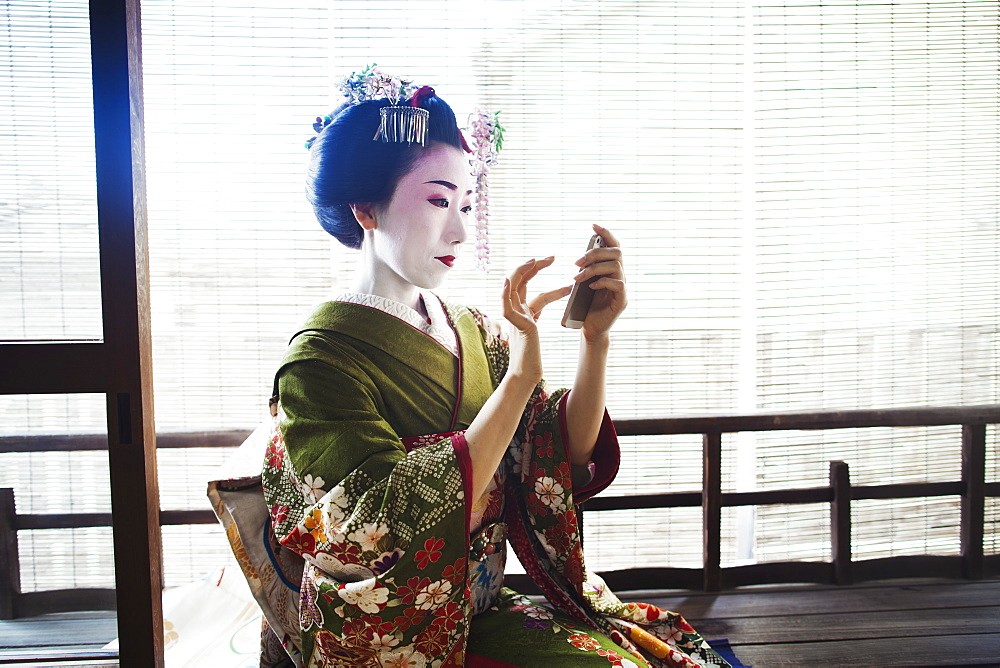 A woman dressed in the traditional geisha style, wearing a kimono and obi, with an elaborate hairstyle and floral hair clips, with white face makeup with bright red lips and dark eyes taking a selfie of herself, Japan