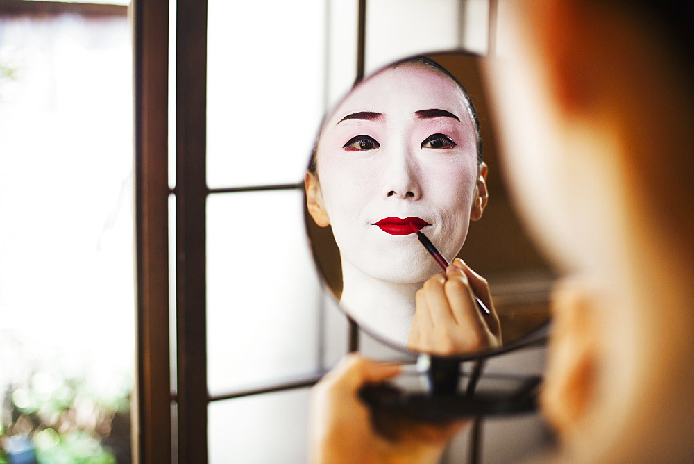 Geisha woman with traditional white face makeup applying bright red lipstick with a brush, using a mirror, Japan