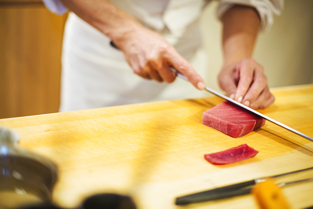 A chef working in a small commercial kitchen, an itamae or master chef slicing fish with a large knife for making sushi, Japan