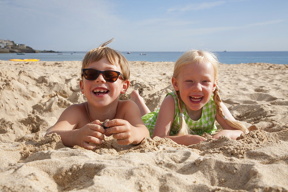 A boy and girl lying on their stomachs on the sand, laughing and looking at the camera, England