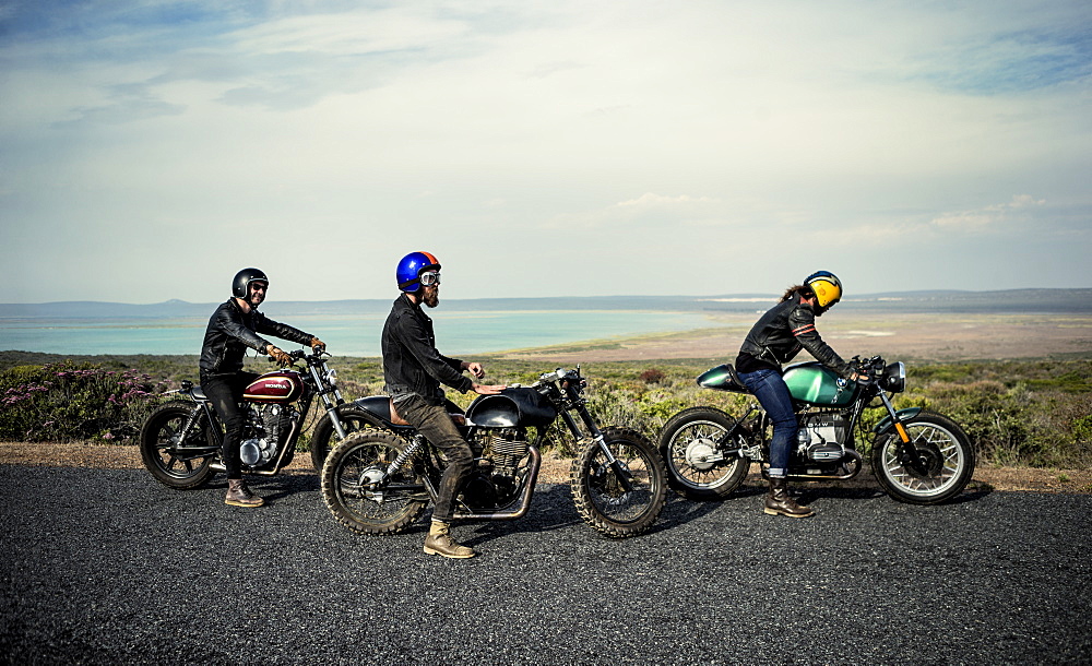 Three men wearing open face crash helmets sitting on cafe racer motorcycles on a rural road, United States of America