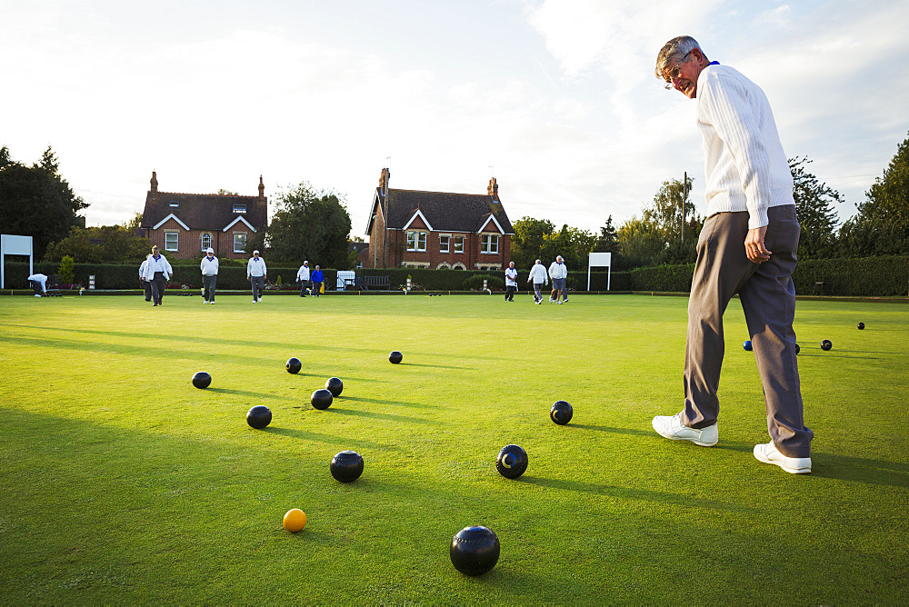 A lawn bowls player walking across the green, the playing surface, at an end with bowls clustered around the yellow jack ball, England, United Kingdom