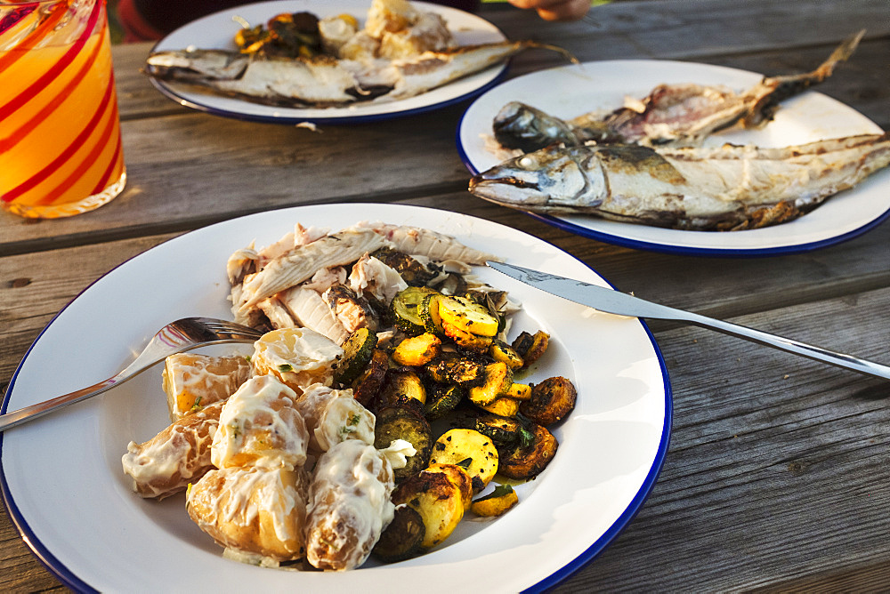 A meal outdoors at a wooden table, plates with grilled mackerel fish, vegetables and potatoes. A drinking glass, England, United Kingdom