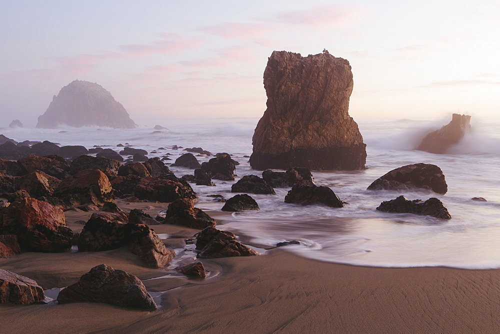 Seascape with breaking waves on rocks on sandy beach at dusk.