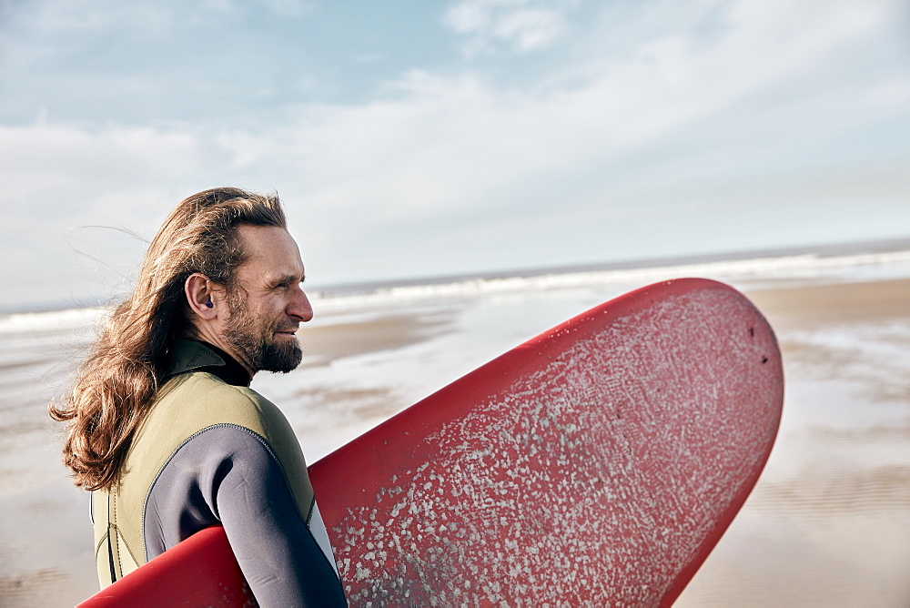 Man holding a full size surf board standing on beach looking out to sea, United Kingdom