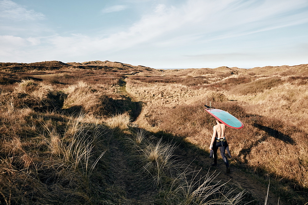 Man walking on a path in the sand dunes towards sea with surf board balancing on his head, United Kingdom