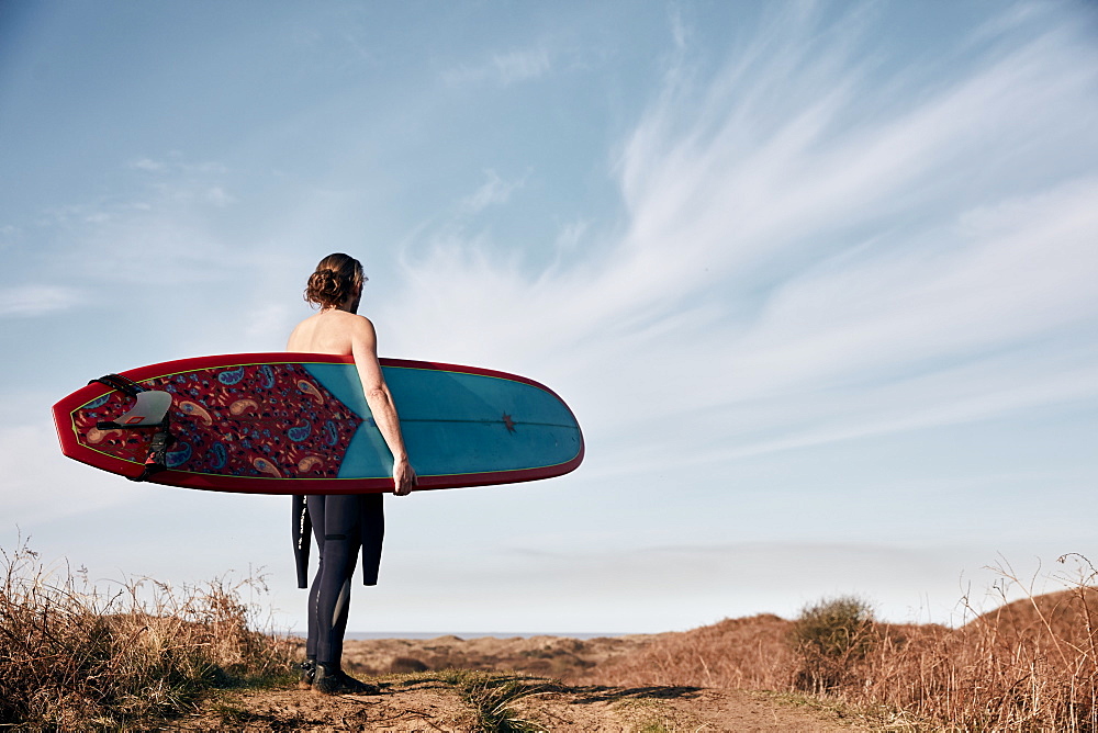 Man walking on a path in the sand dunes towards sea carrying a surf board, United Kingdom
