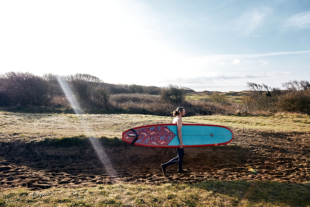 Man holding surf board walking along sandy path towards the sea, United Kingdom