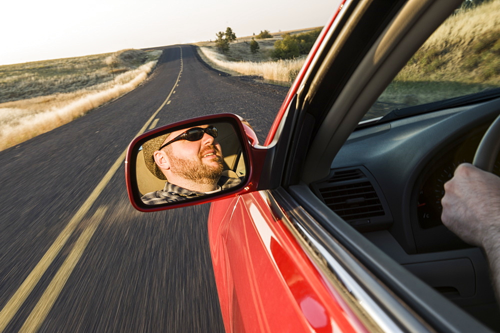 View in the rear view drivers mirror of a Caucasian male driving a car on a road trip in eastern Washington State, United States of America