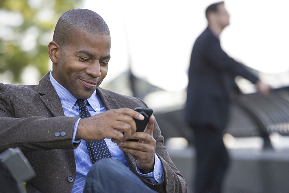 Business people in the city. Keeping in touch on the move. A man seated using his smart phone. A man in the background, New York City, USA