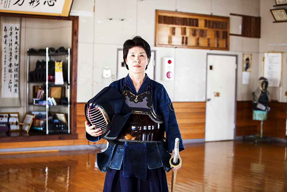 Female Japanese Kendo fighter standing in a gym, holding Kendo mask and sword, looking at camera, Kyushu, Japan