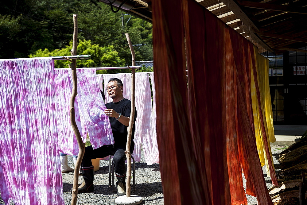 Japanese man sitting outside a textile plant dye workshop, hanging up freshly dyed pink fabric, Kyushu, Japan