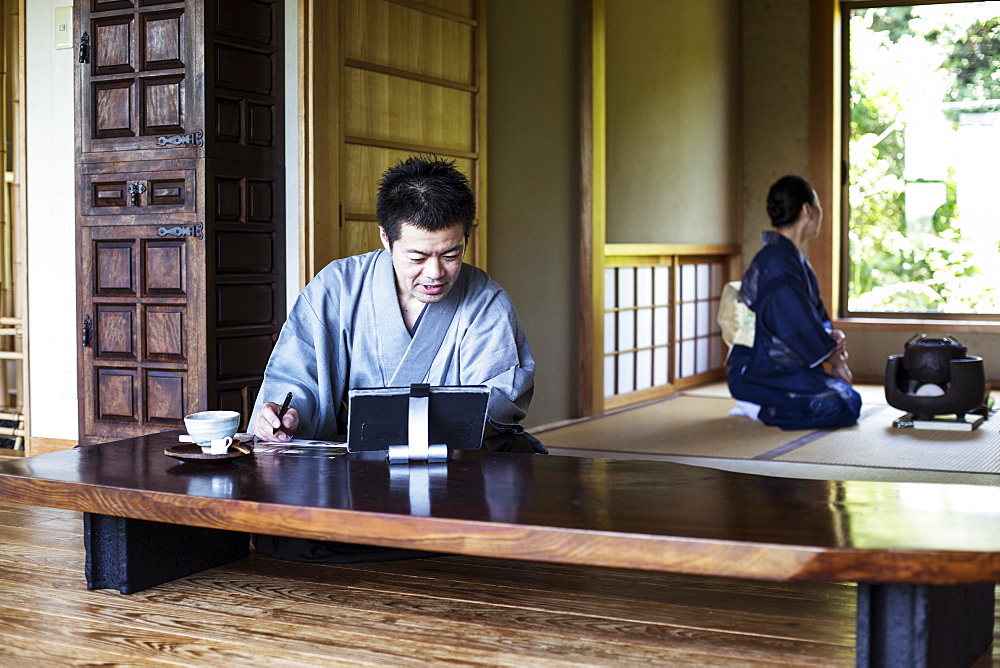 Japanese man wearing kimono sitting on floor in traditional Japanese house, looking at digital tablet, Kyushu, Japan