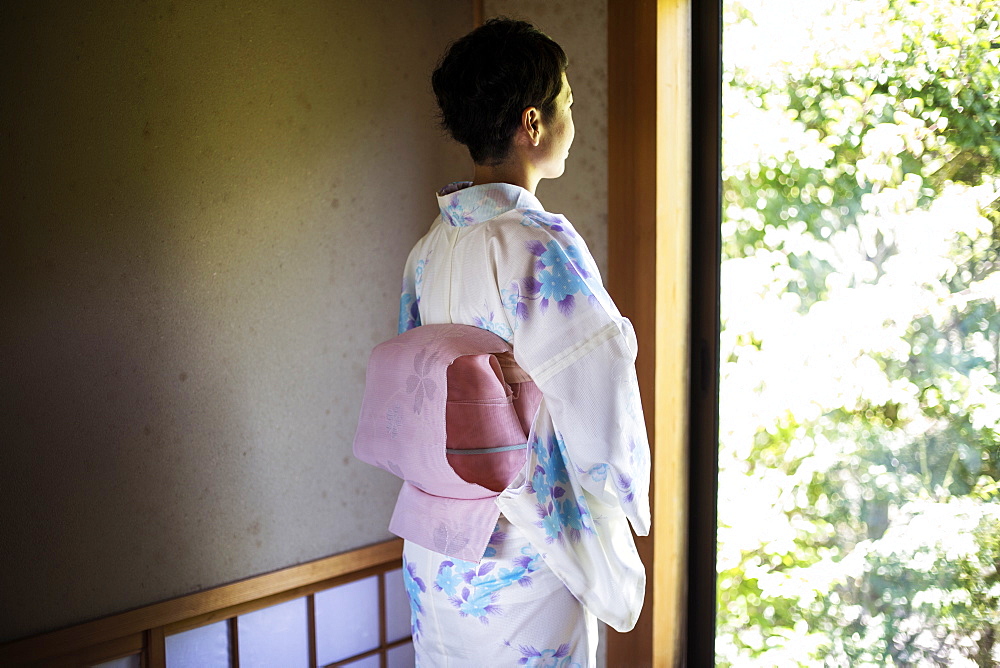 Rear view of Japanese woman standing at window, wearing traditional white kimono with blue floral pattern and pink obi, Kyushu, Japan