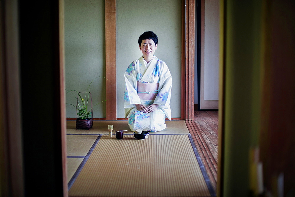 Japanese woman wearing traditional white kimono with blue floral pattern kneeling on tatami mat during tea ceremony, Kyushu, Japan