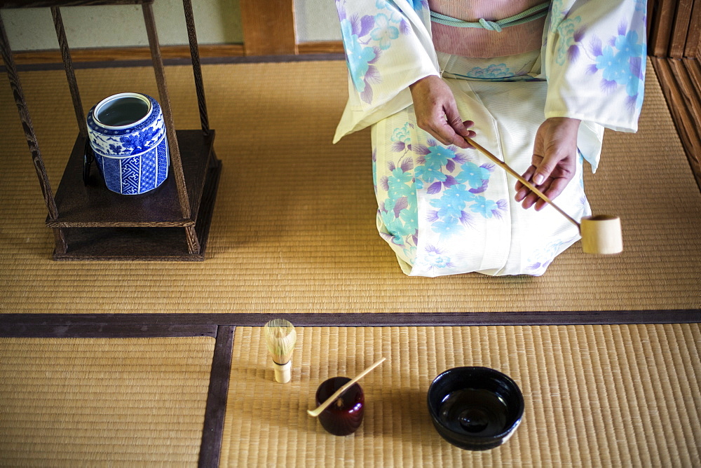 High angle view of Japanese woman wearing traditional white kimono with blue floral pattern kneeling on tatami mat during tea ceremony, holding a Hishaku, a bamboo ladle, Kyushu, Japan